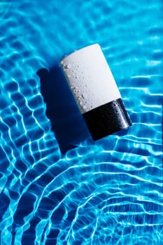 Fashion trendy bottle of perfume toilet water, lotion on blue wave splash background. Beauty cosmetology product. Black and white container making tides. Elegant luxury scent. Vertical. Copy space