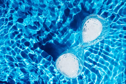 Blue abstract glass hourglass with white sand in water reflect surface pool. Renovation of time. Skin body care, anti age retirement concept. Moisturing dehydrated process. Deadline time. Flat lay.