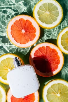 White square perfume bottle on green blue transparent reflect rippled wave water surface, lemon and grapefruit citrus slices. Fashion dermatology skin care. Vertical. Top view