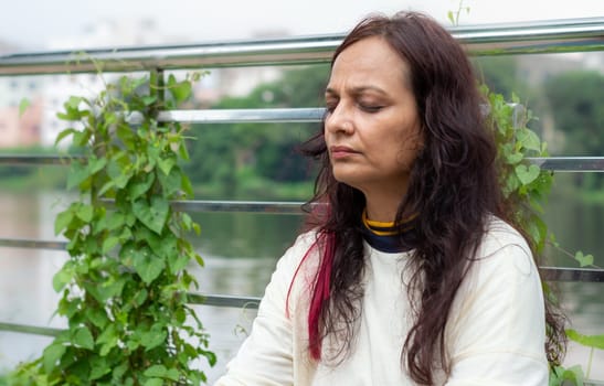 Active Mid Adult Woman in meditation sitting with eyes closed concentrate in a public park against garden leaves in the background. Front view. Head and Shoulder Shot. Close up Portrait.