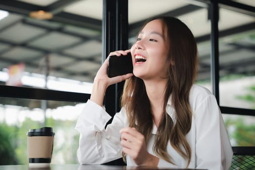Happy female lifestyle sitting in cafe near window she cellphone on mobile phone, Asian young business woman freelance holding smartphone and coffee paper cup at coffee shop