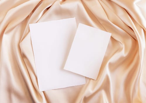 Blank cards on beige silky fabric drapery top view, mockup. Romantic scene with vertical paper cards, greeting or wedding Invitation. .