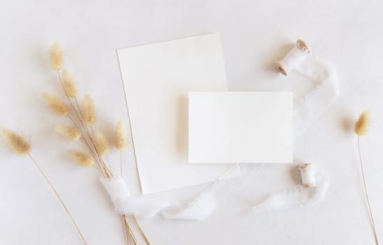 Blank cards near silk ribbons and dried hare's tail grass top view on white, mockup. Boho romantic scene with vertical cards. Bohemian or close to nature greeting or wedding Invitation