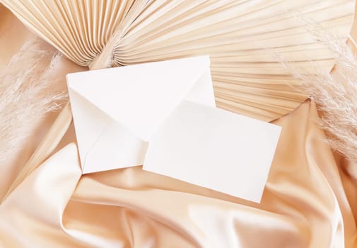 Card and envelope on beige satin fabric and dried palm leaf close up, mockup. Romantic Boho scene with horizontal paper card and envelope. Bohemian or close to nature greeting or wedding Invitation.