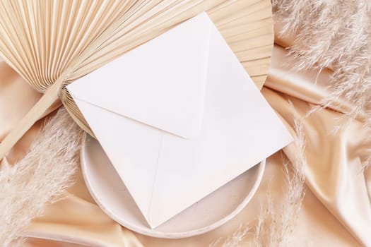 Square envelope on plate with dried palm leaf and beige silky fabric close up, mockup. Romantic scene with blank envelope. Bohemian pastel greeting or wedding Invitation .