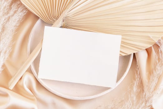 Blank card on plate with dried palm leaf and beige silky fabric close up, mockup. Romantic scene with horizontal paper card. Bohemian pastel greeting or wedding Invitation .