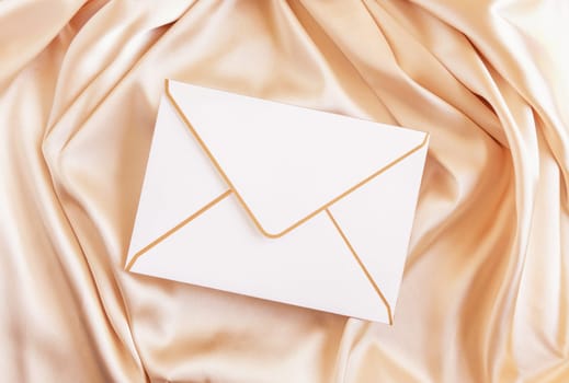 Blank envelope on beige silky fabric drapery top view, mockup. Romantic scene with white envelope, greeting or wedding Invitation.