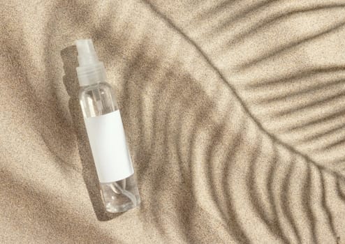 Cosmetic spray bottle with blank label on beige sand top view. Packaging mockup. Natural skincare product for beauty routine. Summer sunny day with palm leaf hard shadow, tropical vacation mood