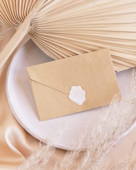 Sealed envelope on plate with dried palm leaf and beige silky fabric close up, mockup. Romantic scene with kraft paper envelope. Bohemian pastel greeting or wedding Invitation .