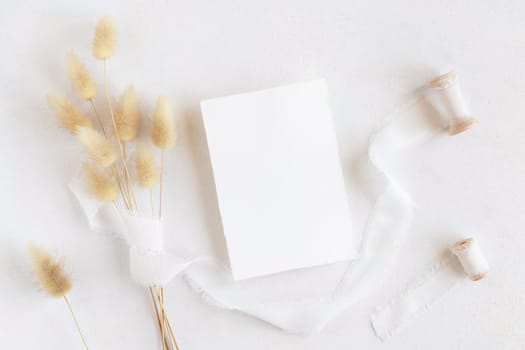 Blank card near silk ribbons and dried hare's tail grass top view on white, mockup. Boho romantic scene with vertical card. Bohemian or close to nature greeting or wedding Invitation