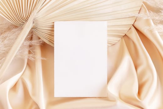 Blank card on beige satin fabric and dried palm leaf close up, mockup. Boho scene with vertical paper card. Bohemian or close to nature greeting or wedding Invitation. Romantic composition.