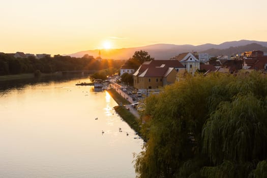 Flowing river with old houses in Maribor in sunset. Lake in city lined with historical bouildings in Slovenia. Touristic destination with water in golden hour.