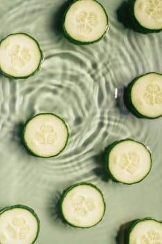 Cucumber slices lie on surface of rippled transparent fresh green exhilarant water gel with flecks, waves, shadow. Expanding circles on surface from fallen drop. Health care. Copy space. Vertical