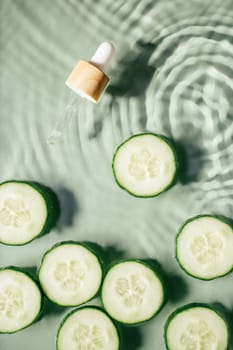 Facial pipette serum, whey beauty product near cucumber slices in water splashing fresh transparent, flecks. Flat lay, top view. Summer beach. Organic skin face and body moisturizing. Vertical