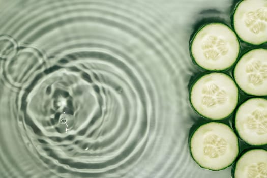 Cucumber slices lie on surface of rippled transparent fresh green exhilarant water gel with flecks, waves, shadow. Concentric expanding circles on surface from fallen drop. Health care. Copy space