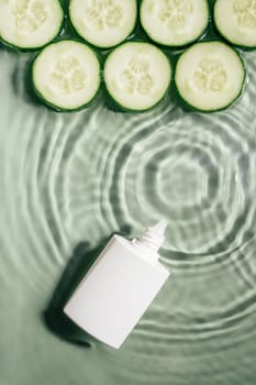 White beauty product cream vial tube bottle with rows of cucumber slices on surface of rippled transparent green water with fleck, wave, shadow. Concentric expanding circles from fallen drop. Vertical