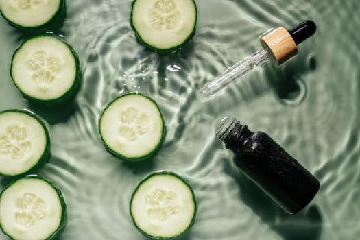 Facial pipette serum black bottle, whey beauty product near cucumber slices in water splashing fresh transparent, flecks. Flat lay, top view. Organic skin face and body moisturizing and rejuvenating