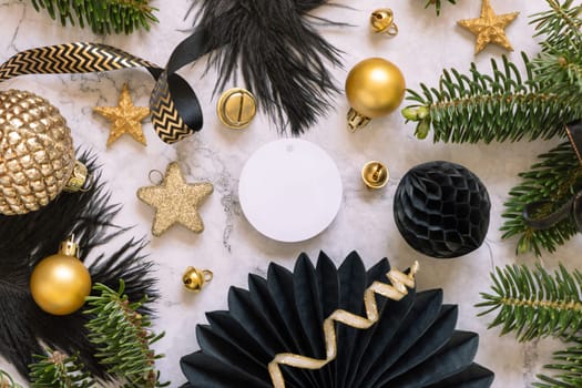 Paper round gift tag between fir tree branches and black and golden Christmas decorations on a white marble table top view. Mockup, copy space. Winter holiday flat lay with blank label