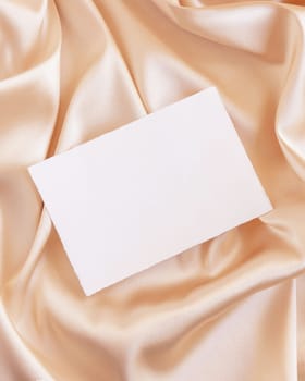 Blank card on beige silky fabric drapery top view, mockup. Romantic scene with horizontal paper card, greeting or wedding Invitation. .