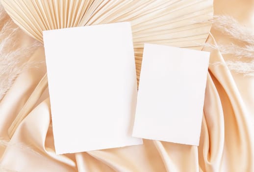 Blank cards on beige satin fabric and dried palm leaf close up, mockup. Boho scene with vertical paper cards. Bohemian or close to nature greeting or wedding Invitation. Romantic composition.