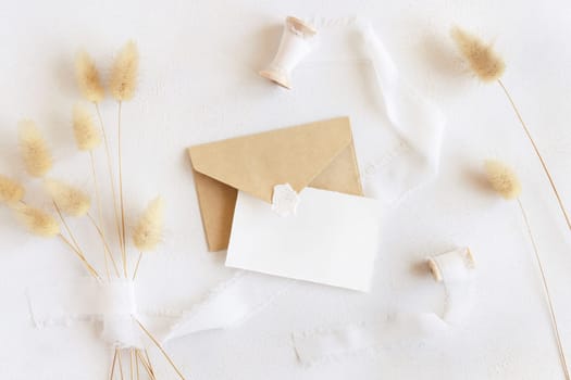 Blank card and envelope near silk ribbons and dried hare's tail grass top view on white, mockup. Boho romantic scene with horizontal cards. Bohemian or close to nature greeting or wedding Invitation