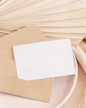 Blank card and envelope on plate with dried palm leaf and beige silky fabric close up, mockup. Romantic scene with horizontal paper card. Pastel greeting or wedding stationery.