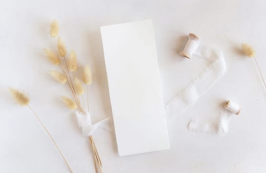 Blank cards near silk ribbons and dried hare's tail grass top view on white, mockup. Boho romantic scene with vertical card. Bohemian or close to nature greeting or wedding Invitation