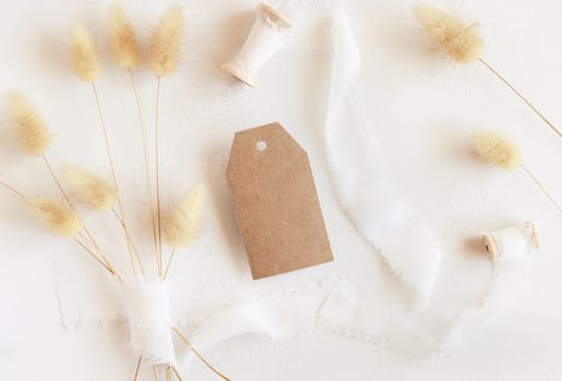 Blank gift tag near silk ribbons and dried hare's tail grass top view on white, mockup. Boho romantic scene with kraft paper gift tag. Bohemian or close to nature greeting