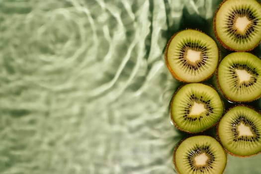 Kiwi slices lie on surface of rippled transparent fresh green exhilarant water gel with flecks, waves, shadow. Concentric expanding circles on surface from fallen drop. Health care. Copy space