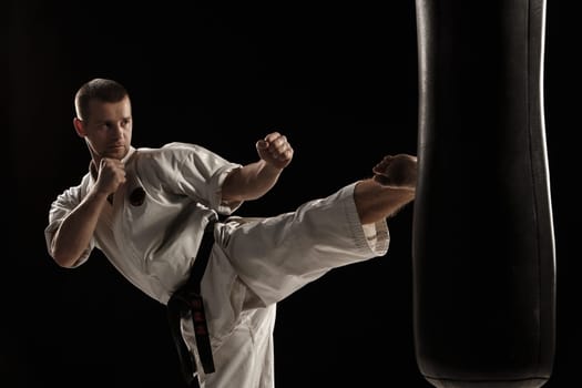 Man in white kimono training karate round kick in a punching bag over black background