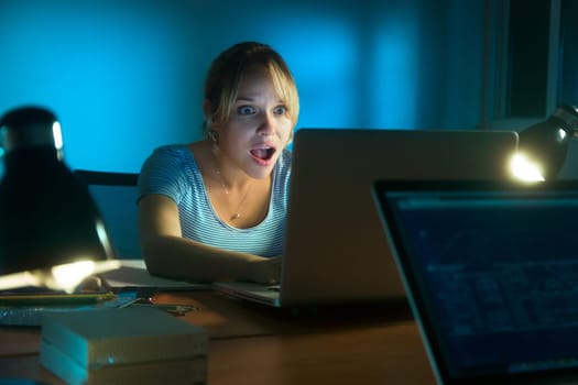 Beautiful woman working late at night in office, surfing the web and reading a post on social network with laptop computer. The girl looks at the screen with open mouth