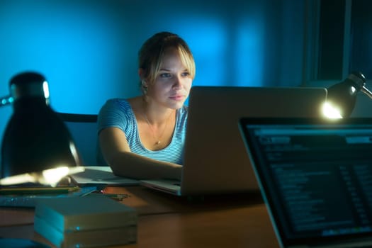 Beautiful woman working late at night in office, surfing the web and writing post on social network with laptop computer. The girl is concentrated and serious
