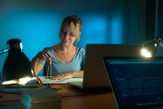 Beautiful woman working as interior designer, staying late at night in office with drawings and laptop computer to complete a project. The girl draws a circle with compasses