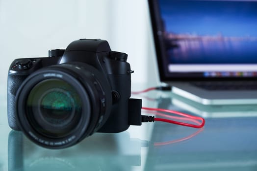 Still life of dslr camera connected with USB cable to laptop computer on glass table. The photo camera is transferring images to PC in background. Copy space