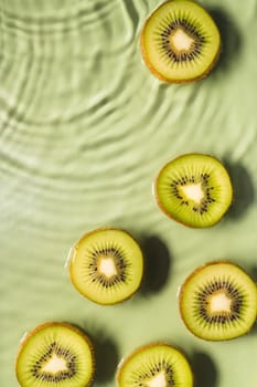 Kiwi slices lie on surface of rippled transparent fresh green exhilarant water gel with flecks, waves, shadow. Concentric expanding circles on surface from fallen drop. Vertical. Copy space