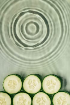 Cucumber slices lie on surface of rippled transparent fresh green exhilarant water gel with flecks, waves, shadow. Expanding circles on surface from fallen drop. Health care. Copy space. Vertical