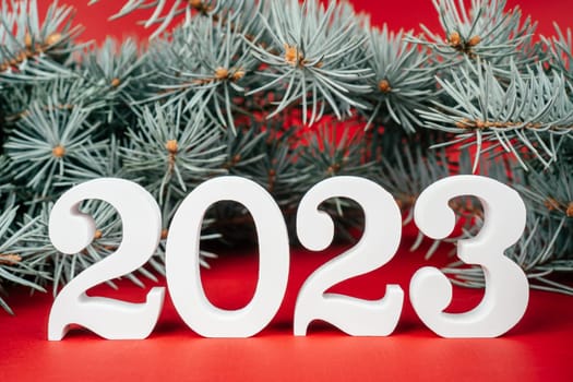 Happy New Year 2023. White wooden numbers 2023 stand with lush green Christmas tree branches on red background. Merry Christmas.