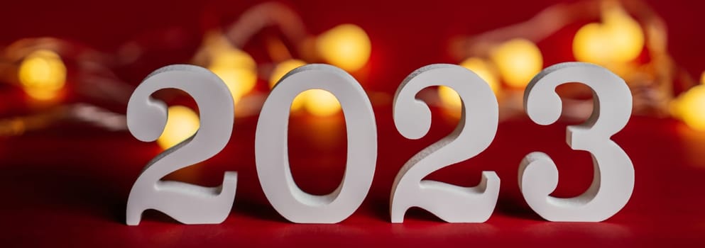 Happy New Year 2023. White wooden numbers 2023 stand on red background with garland light bulbs. Merry Christmas.