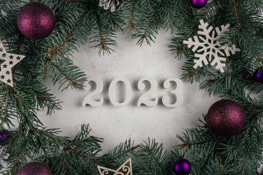 White wooden numbers 2023 lying on white concrete surface with branches of Christmas tree, Christmas tree decorations and purple balls. Merry Christmas and Happy New Year 2023. Top view