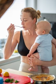 Woman cooking and tasing food while holding four months old baby boy in her hands.