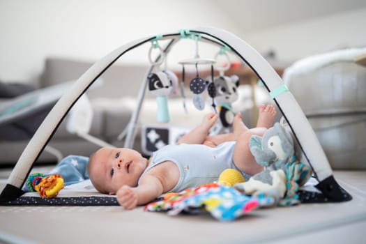 Cute baby boy playing with hanging toys arch on mat at home Baby activity and play center for early infant development. Baby playing at home.