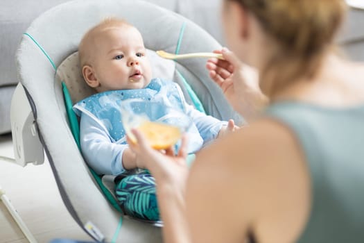 Mother spoon feeding her baby boy child in baby chair with fruit puree. Baby solid food introduction concept