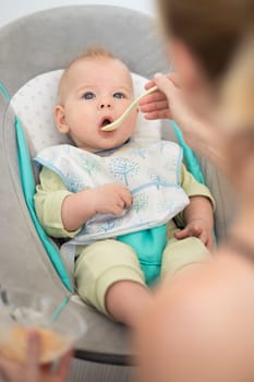 Mother spoon feeding her baby boy child in baby chair with fruit puree. Baby solid food introduction concept