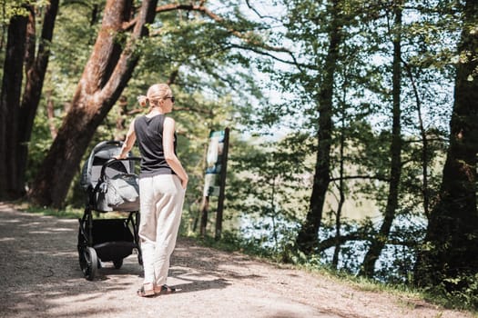 Rear view of casualy dressed mother walking with baby stroller in city park. Family, child and parenthood concept