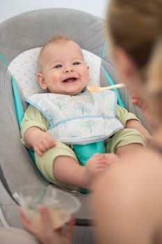 Mother spoon feeding her baby boy child in baby chair with fruit puree. Baby solid food introduction concept