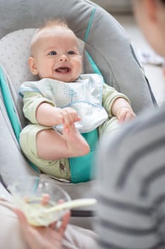 Mother spoon feeding her baby boy child in baby chair with fruit puree. Baby solid food introduction concept