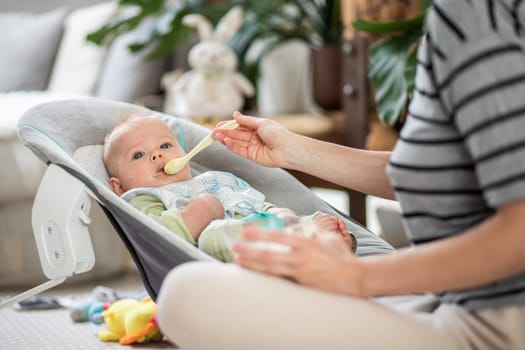 Mother spoon feeding her baby boy child in baby chair with fruit puree. Baby solid food introduction concept
