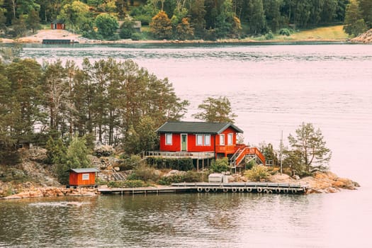 Sweden. Many Beautiful Red Swedish Wooden Log Cabin House On Rocky Island Coast In Summer. Lake Or River Landscape.