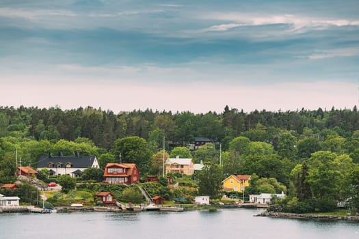 Sweden. Many Beautiful Red Swedish Wooden Log Cabins Houses On Rocky Island Coast In Summer Sunny Evening. Lake Or River Landscape.