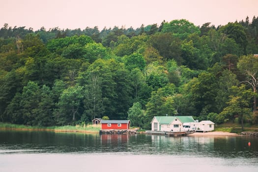 Sweden. Beautiful Red Swedish Wooden Log Cabin House On Rocky Island Coast In Summer. Lake Or River And Forest Landscape.
